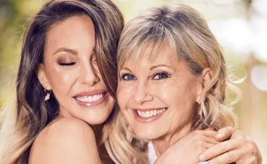 Two women smiling warmly, embracing each other, with a blurred outdoor background.