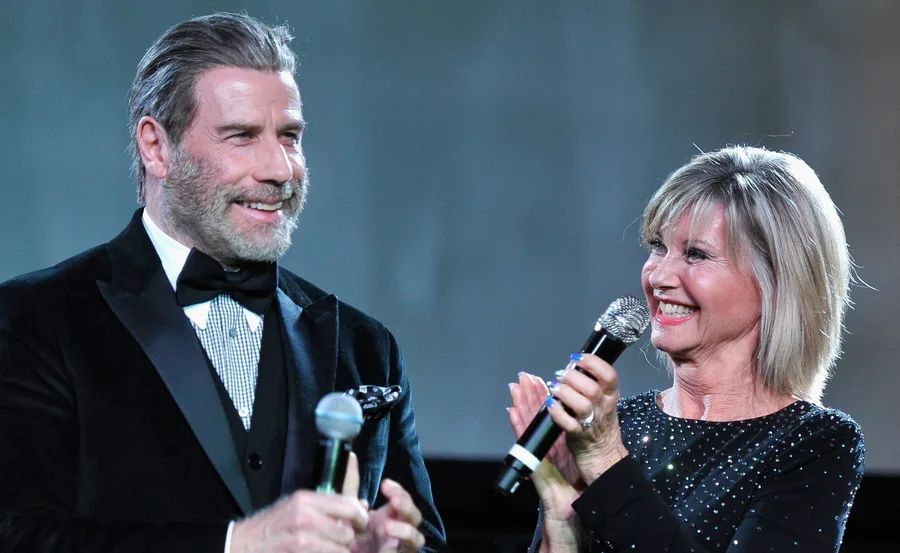 John Travolta and Olivia Newton-John smiling and holding microphones on stage.