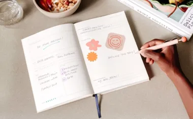 Person writing in a journal with stickers, next to a bowl of granola and yogurt on a table.