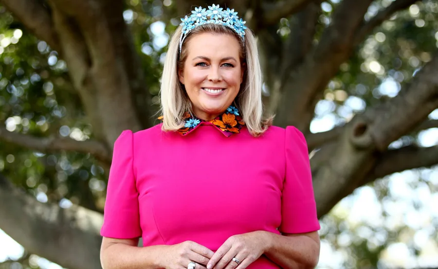 Woman in a pink dress and blue flower headband smiling in front of a tree.