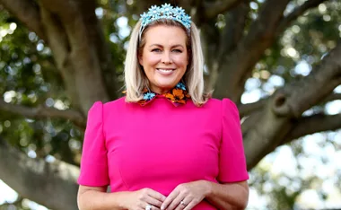 Woman in a pink dress and blue flower headband smiling in front of a tree.