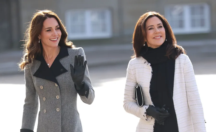 Two women walking outdoors, smiling, one waves her hand, both dressed in winter coats and gloves.