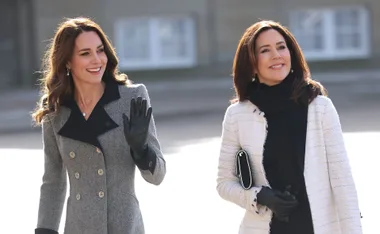 Two women walking outdoors, smiling, one waves her hand, both dressed in winter coats and gloves.