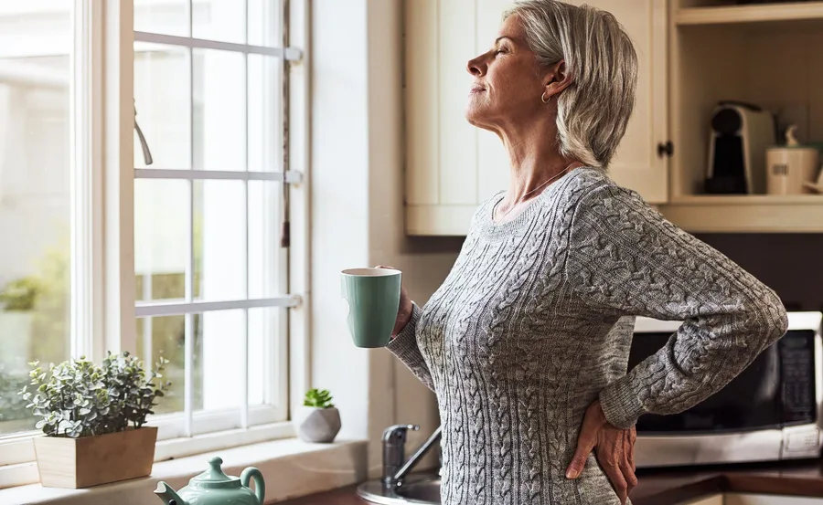 Elderly woman in a kitchen looking out the window, holding a green mug and wearing a gray sweater.