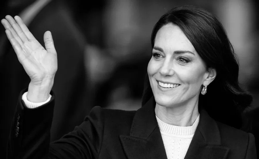 A woman smiling and waving, wearing a dark coat with earrings, in a black and white photo.