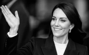 A woman smiling and waving, wearing a dark coat with earrings, in a black and white photo.