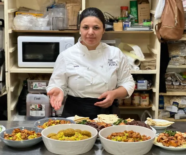 Chef in a kitchen with various dishes, wearing a white chef's jacket with "The Ezidi Place" logo.
