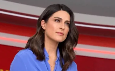A person in a blue shirt with wavy dark hair, sitting in a studio with red and grey background.