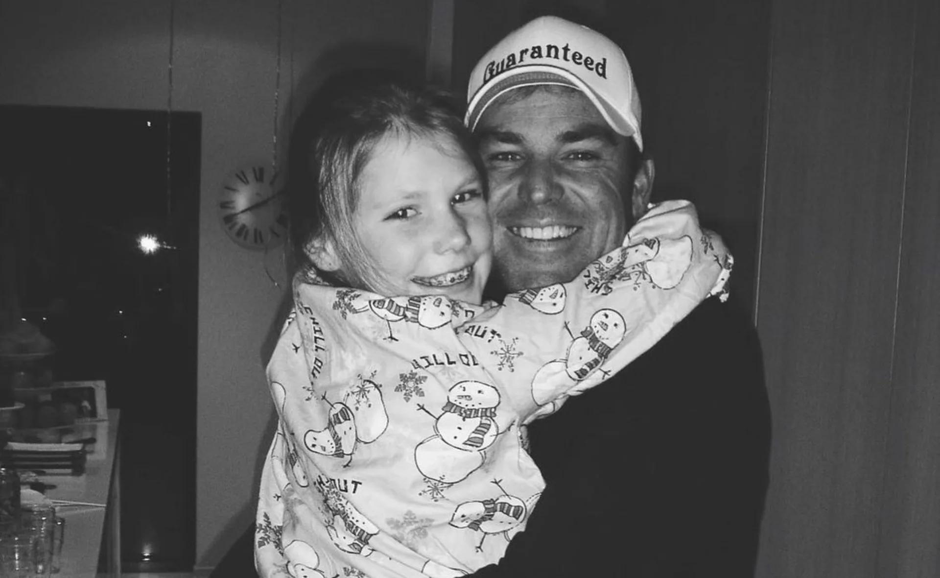 A man in a "Guaranteed" hat hugs a smiling child wearing snowman pajamas in a cozy room.