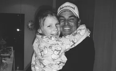 A man in a "Guaranteed" hat hugs a smiling child wearing snowman pajamas in a cozy room.