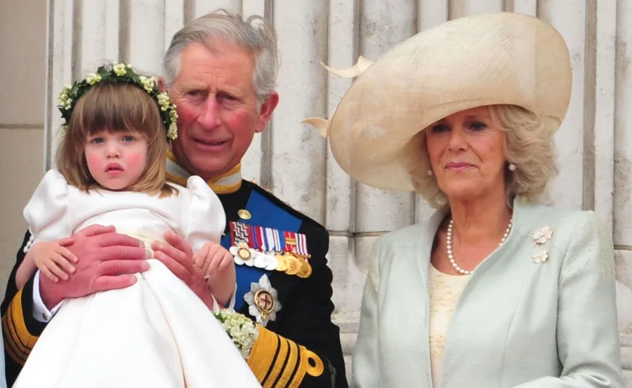 A man in uniform holds a young girl in a white dress, standing next to a woman in a large hat.
