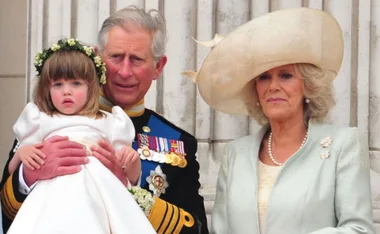 A man in uniform holds a young girl in a white dress, standing next to a woman in a large hat.