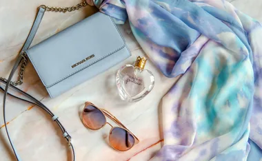 Michael Kors purse, Vera Wang Princess perfume, sunglasses, and a watercolor-patterned scarf on marble background.