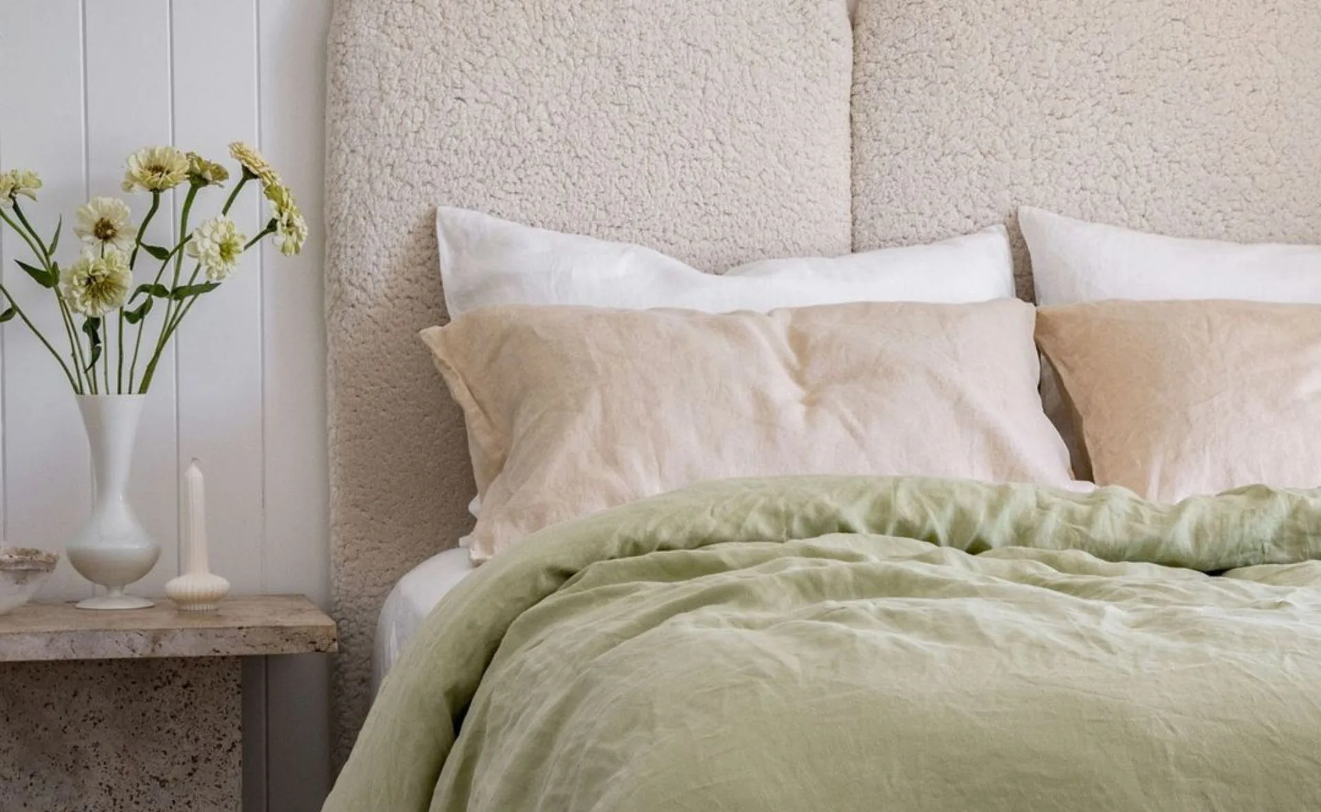 Cozy bedroom scene with beige pillows, a green duvet, and a vase of flowers on a bedside table.