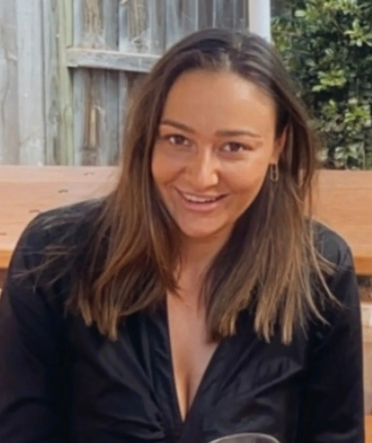 A woman with long hair smiling, seated outdoors with a wooden fence and greenery in the background.