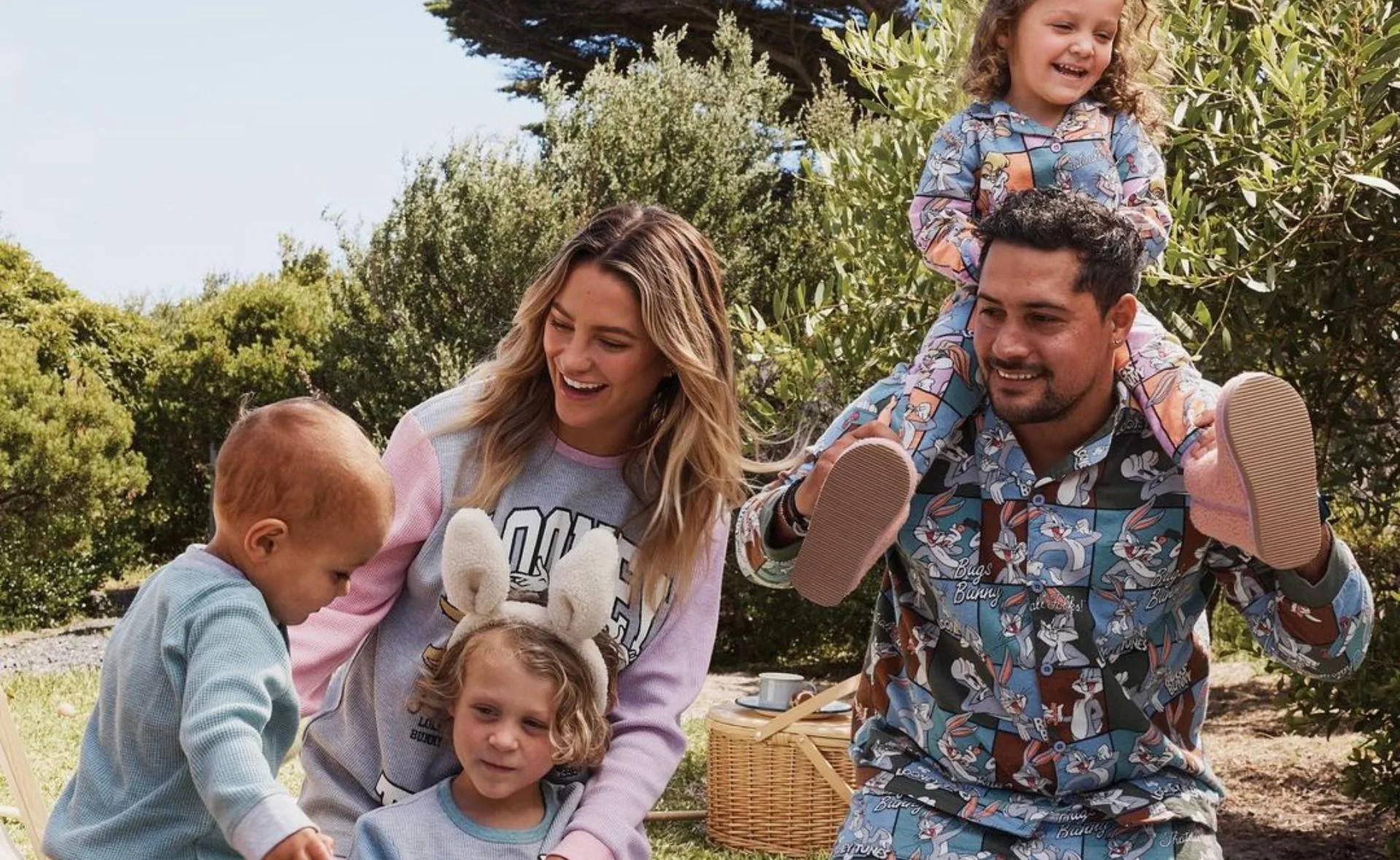 Family wearing matching Bugs Bunny pajamas outdoors, with a child on an adult's shoulders and others playing nearby.