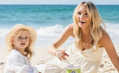 A woman and a young girl wearing a hat smile while sitting on a sunny beach near the ocean.