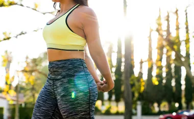 Woman in a yellow sports bra and patterned leggings outdoors, stretching her arms in sunlight.