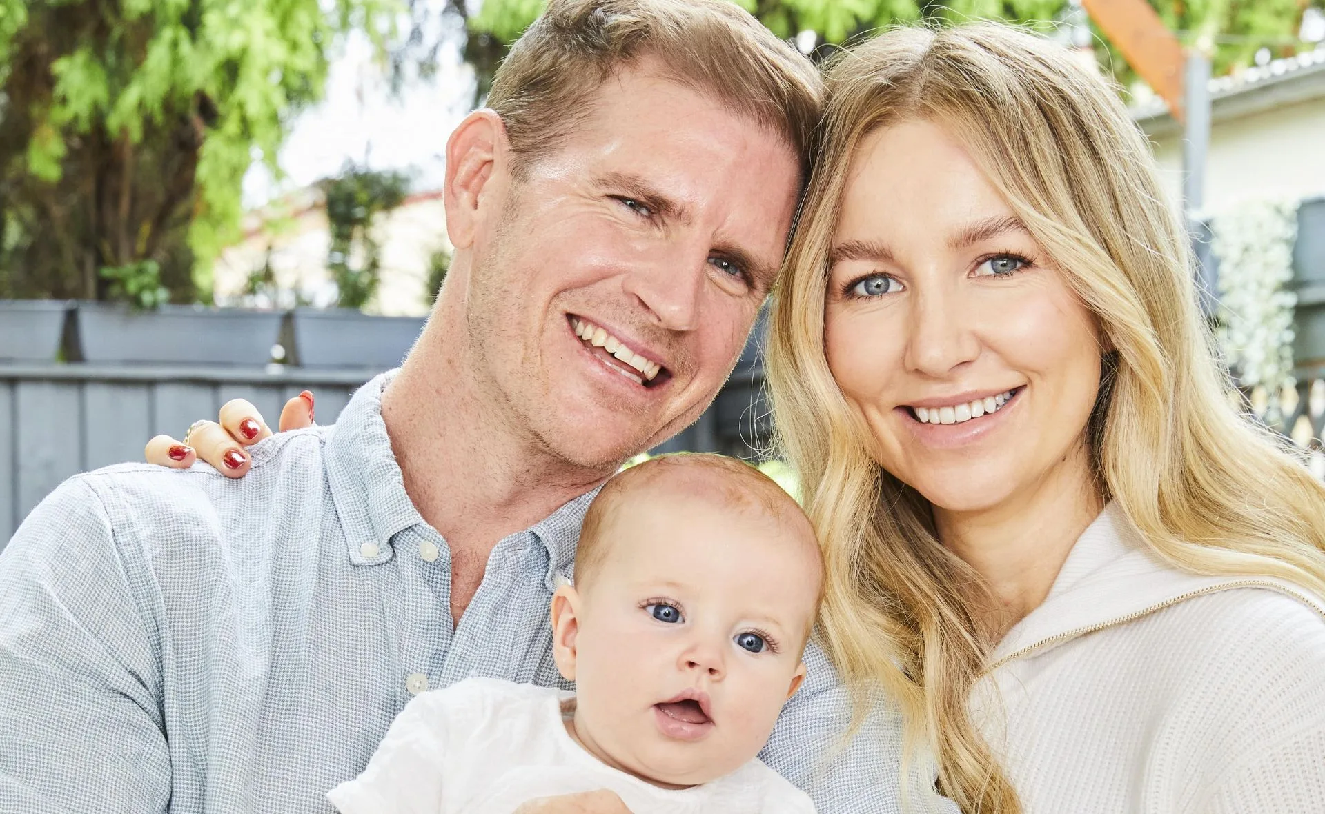 A smiling couple holding a baby, outdoor setting with greenery in the background.