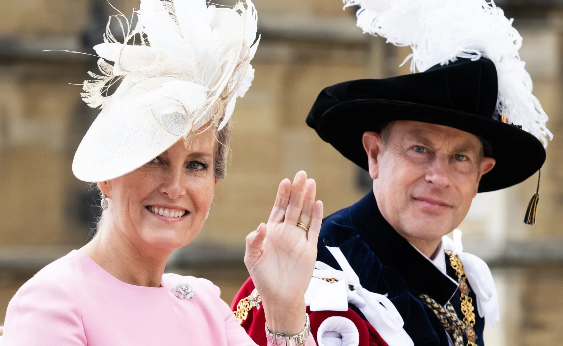 Royal couple, woman in pink with floral hat, man in ceremonial attire with feathered hat, both smiling and waving.