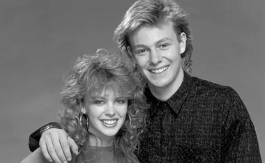 Black and white photo of two smiling actors with 80s hairstyles, likely from the show "Neighbours."