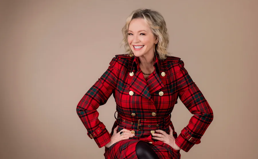 Woman in a red tartan trench coat smiling with hands on hips against a beige background.