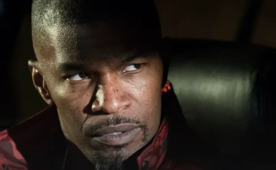 A man in a dark setting with intense expression, wearing a red jacket.