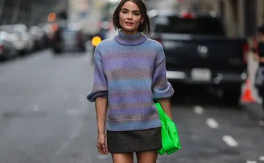 Woman wears a pastel knitted sweater, short skirt, and carries a bright green bag on a city street.