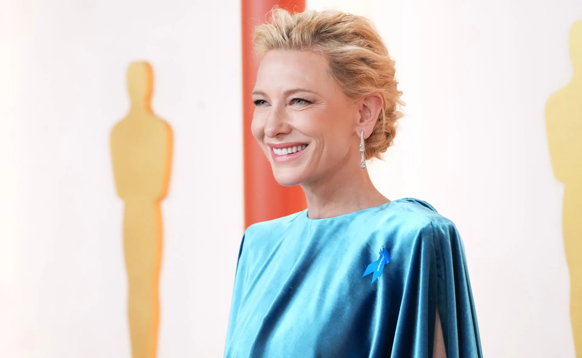 Smiling person in a blue dress with a ribbon, posing on a red carpet with Oscar statuettes in the background.