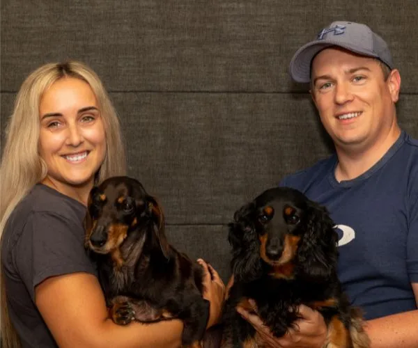 Two people smiling, each holding a dachshund dog against a dark background.
