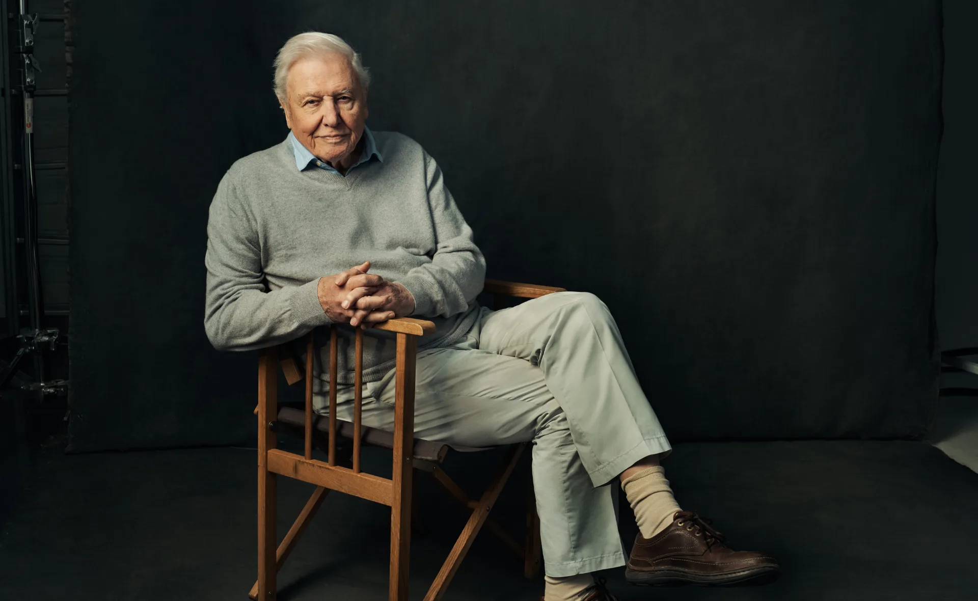 Elderly man in gray sweater and khaki pants sitting on a wooden chair, dark background, looking at the camera.