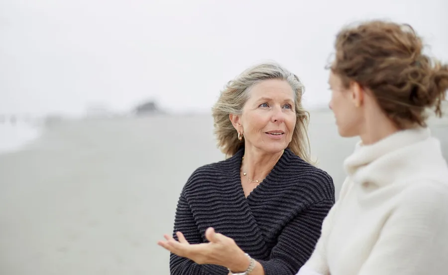 Two women talking on a beach, one with gray hair in a black sweater, the other with brown hair in a white sweater.