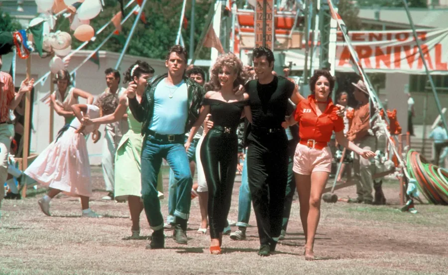 A group of characters from "Grease" walk arm in arm at a carnival, set against a vibrant festive backdrop.