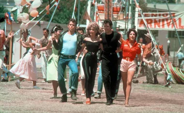 A group of characters from "Grease" walk arm in arm at a carnival, set against a vibrant festive backdrop.