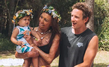 Family smiling outdoors, woman holding child, both wearing flower crowns, man beside them.