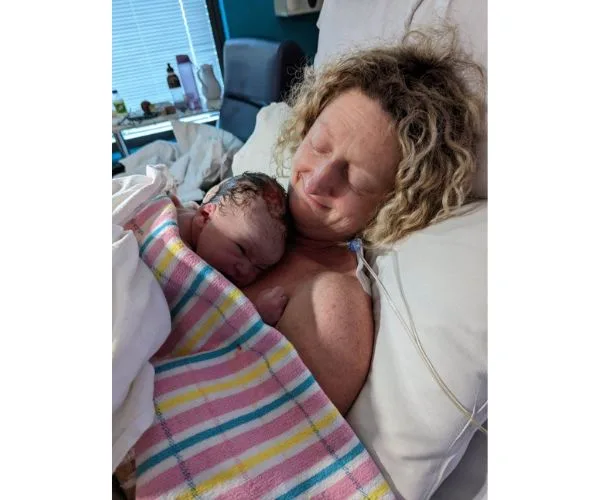 A woman lies in a hospital bed, smiling while holding a newborn baby wrapped in a colorful blanket against her chest.