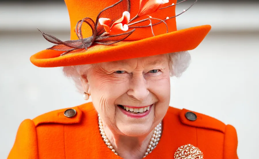 Queen Elizabeth II smiling in a vibrant orange outfit and matching hat adorned with floral accents.