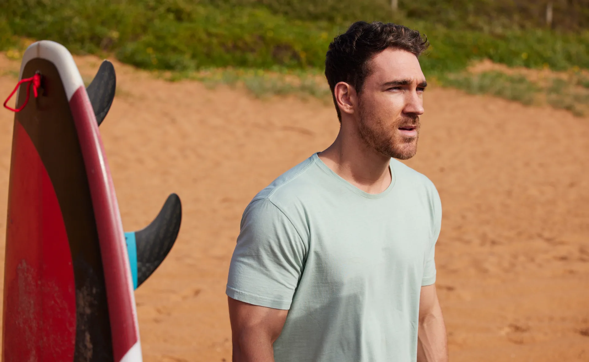Man in a light blue shirt standing on a sandy beach next to a red and white surfboard.