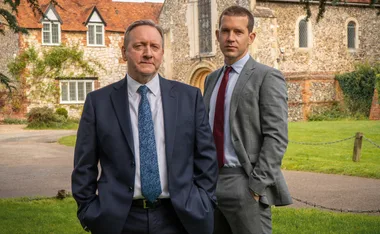 Two men in suits stand in front of a stone building with red roof, surrounded by greenery.