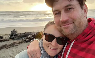 Couple smiling on a beach during sunset, with ocean waves and driftwood in the background.