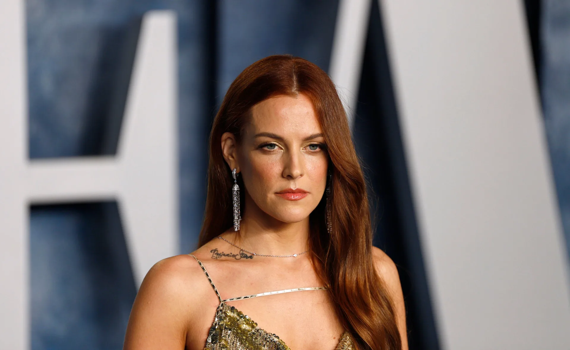 A woman with long red hair, wearing a sequined dress and earrings, stands in front of a blurred background.