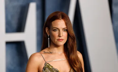 A woman with long red hair, wearing a sequined dress and earrings, stands in front of a blurred background.