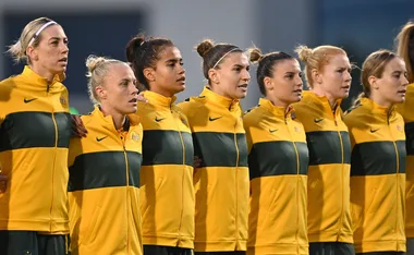 Australian women's soccer team in yellow jackets standing linked before a match.