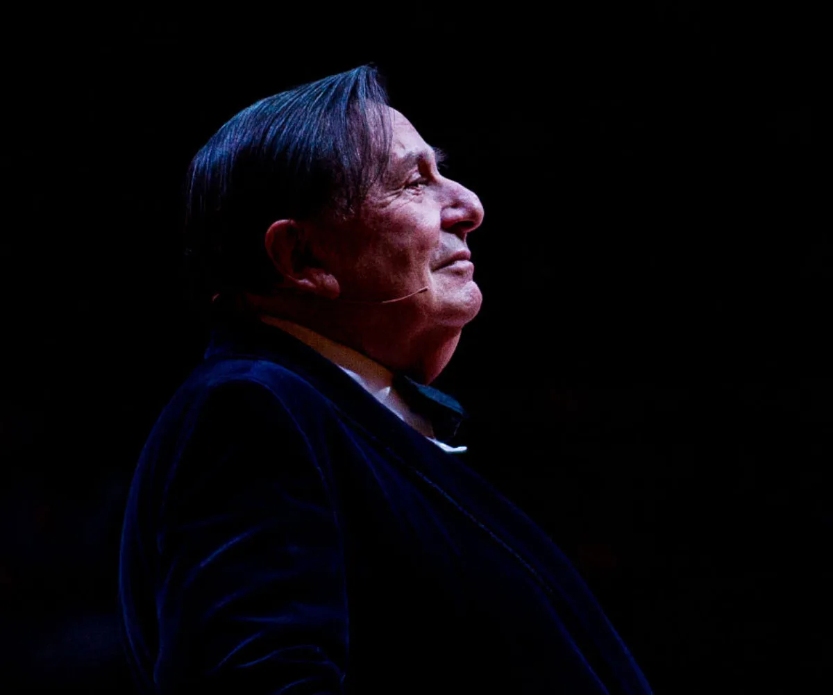 A person in a dark outfit stands in profile onstage, with a microphone, under spotlight against a dark background.