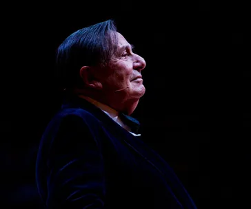 A person in a dark outfit stands in profile onstage, with a microphone, under spotlight against a dark background.