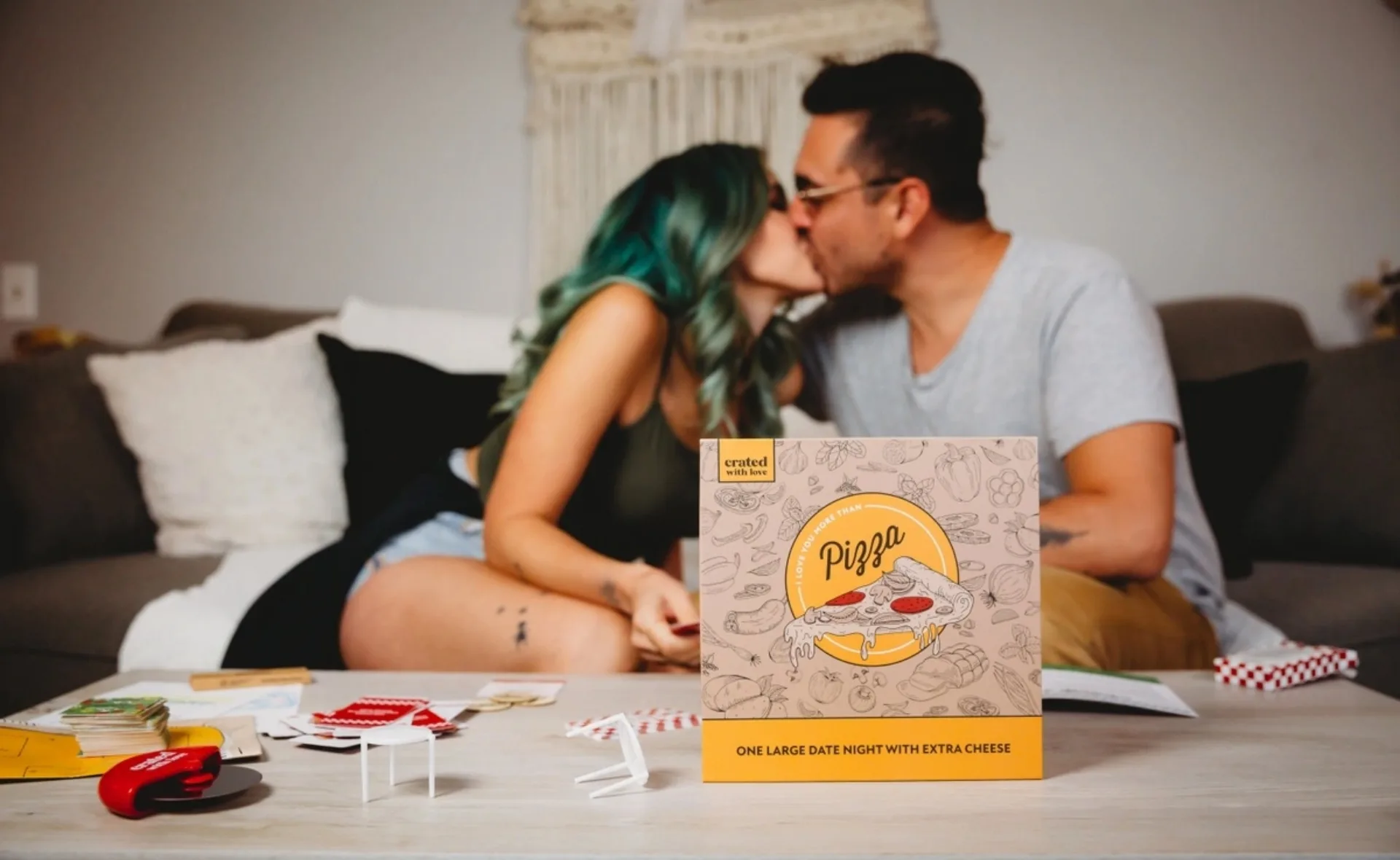 Couple kissing on a couch with a "Crated with Love" date night box featuring a pizza theme in the foreground.