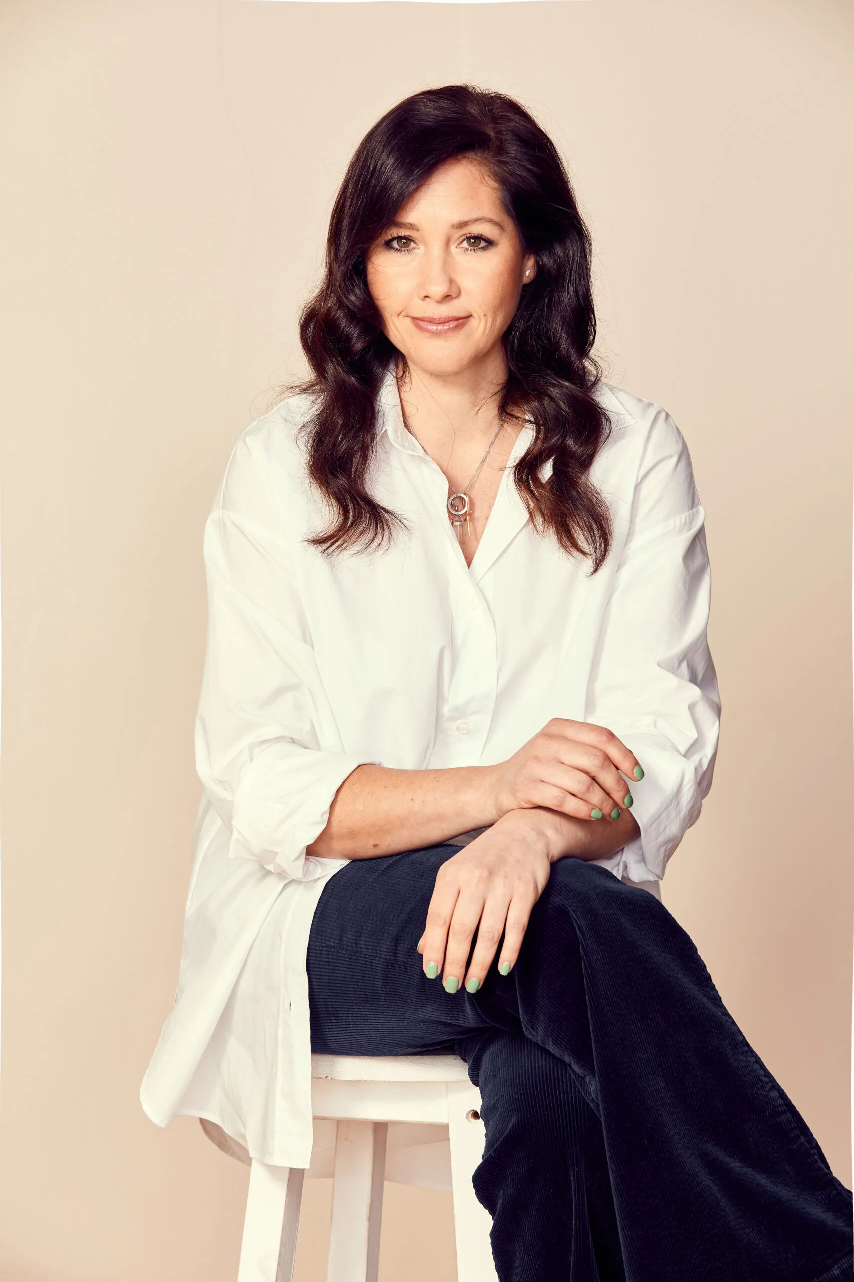 A woman with dark hair, wearing a white shirt, sits on a stool with hands resting on her lap.