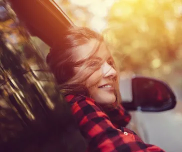 Woman smiling with her head out a car window in autumn sunlight, wearing a red plaid shirt.