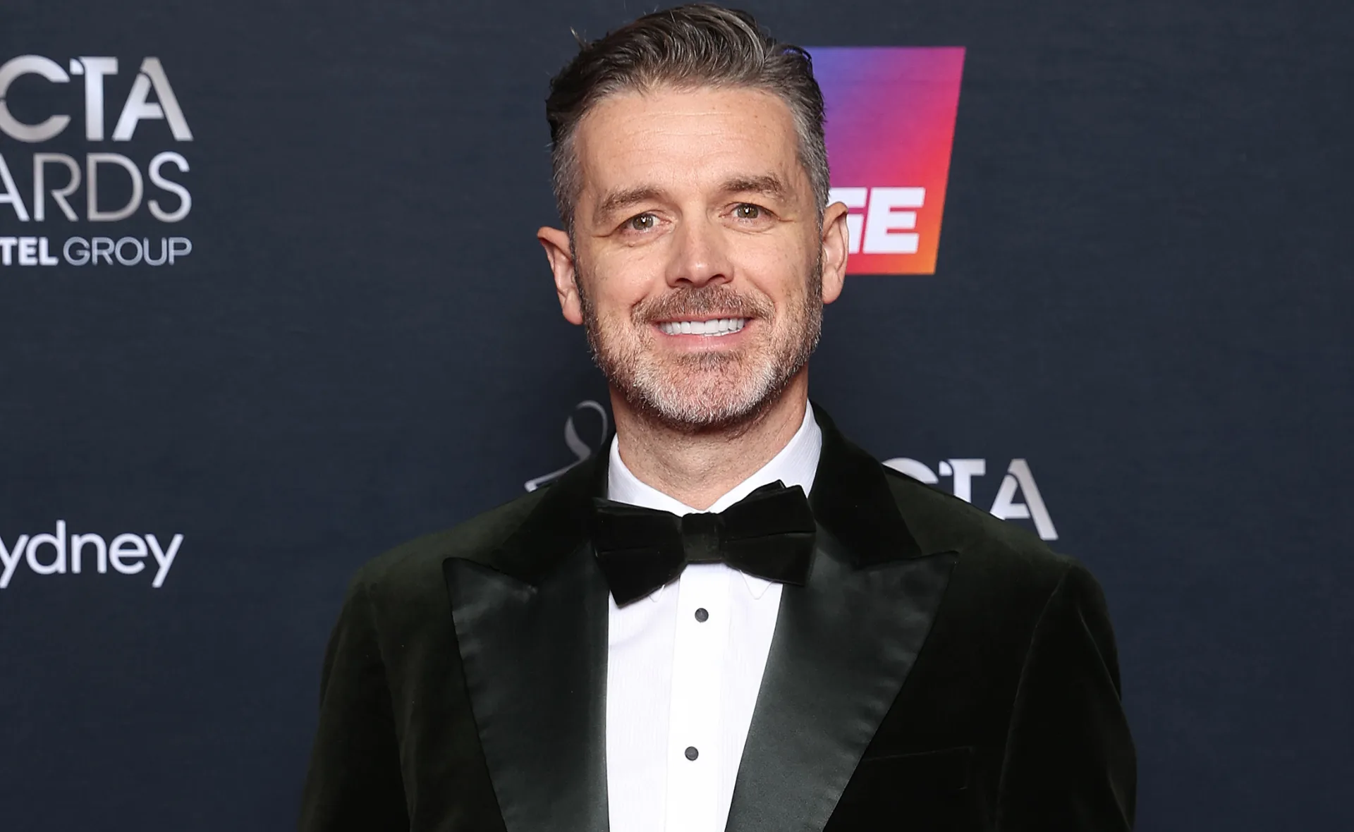 Man in a tuxedo smiling at the AACTA Awards backdrop.