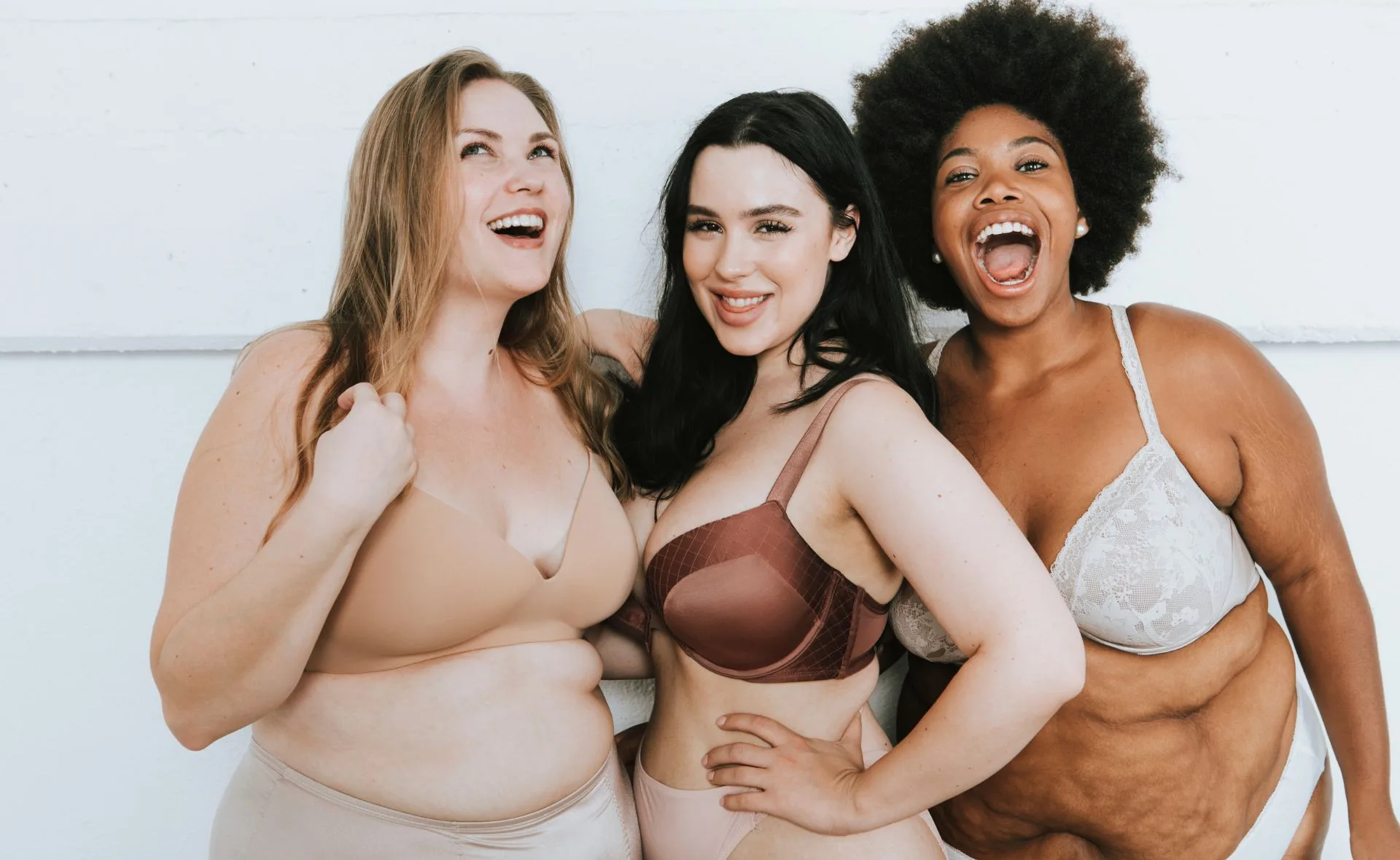 Three smiling women in different styles of bras stand together, displaying body positivity and diversity.